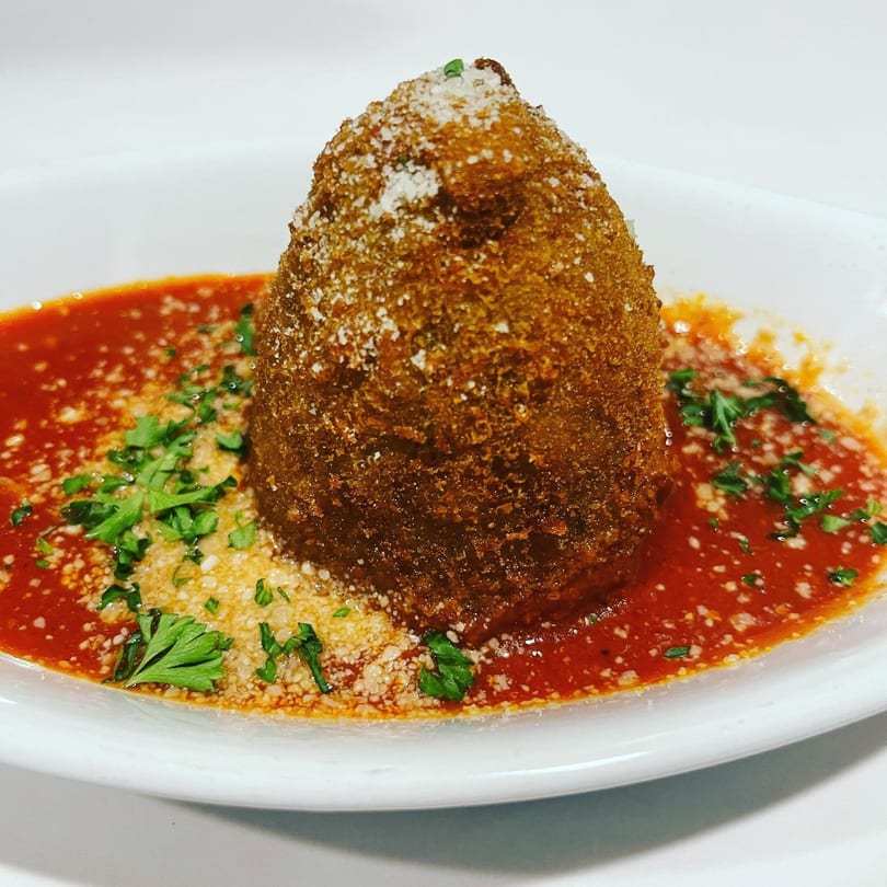 A golden-brown arancini, topped with parmesan and parsley, sits in a pool of red marinara sauce.